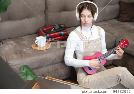 Young caucasian hipster woman is engaged in online ukulele lesson at home, sitting on the floor, on the background of the sofa, in the living room 94282932