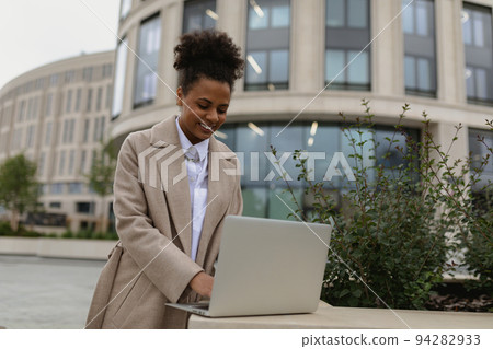 young black woman working on a laptop online against the backdrop of the city with a smile on her 94282933