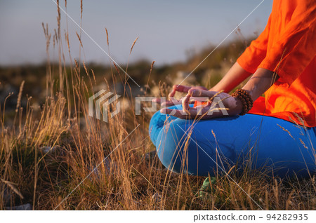 Close-up of a woman and female hands sitting in a lotus position, yoga outdoors in the grass at sunset against the backdrop of a mountain range and clouds, meditation Close-up of a woman and female hands sitting in a lotus position, yoga outdoors in the grass at sunset against the backdrop of a mountain range and clouds, meditation 94282935