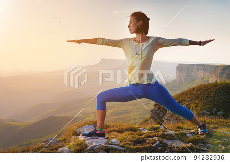 yoga warrior pose of young caucasian woman practicing at sunset in the mountains in the grass. Zen health and wellness concept yoga warrior pose of young caucasian woman practicing at sunset in the mountains in the grass. Zen health and wellness concept 94282936