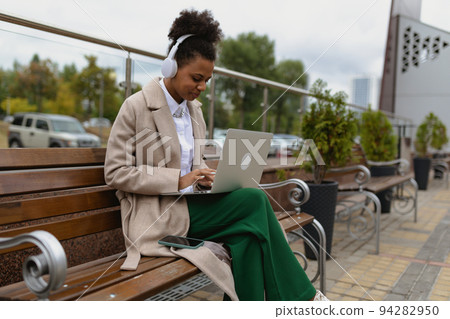 African-American young woman student working online on a laptop with headphones Sitting on a bench African-American young woman student working online on a laptop with headphones Sitting on a bench 94282950