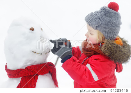 Little boy in red clothes having fun with big snowman. Active winter outdoor leisure for children. Child during stroll in a snowy winter park 94285446