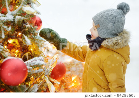 Little boy admiring artificial Christmas tree decorated with gold and red balls and flowers. Traditional city outdoors christmas market. Urban celebrate decoration 94285603