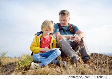Cute schoolchild and mature father hiking together on mountain and exploring nature. Child learning survival skills and orienteering. Concepts of adventure, scouting and hiking tourism for kids. Cute schoolchild and mature father hiking together on mountain and exploring nature. Child learning survival skills and orienteering. Concepts of adventure, scouting and hiking tourism for kids. 94285610
