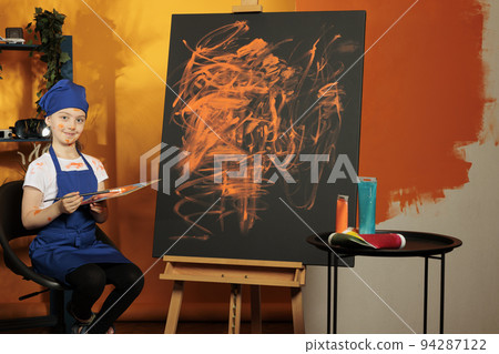 Portrait of small child painting masterpiece with orange color from mixing tray and dye palette, creating art design on canvas with easel. Creative skills to paint colorful artwork with watercolor. Portrait of small child painting masterpiece with orange color from mixing tray and dye palette, creating art design on canvas with easel. Creative skills to paint colorful artwork with watercolor. 94287122