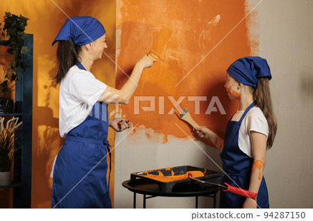 Small family painting walls with orange color paint and brush with bristles, using redecorating tools. Adult and little girl being messy and having fun renovating interior apartment space. Small family painting walls with orange color paint and brush with bristles, using redecorating tools. Adult and little girl being messy and having fun renovating interior apartment space. 94287150