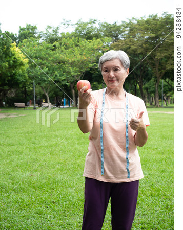 Portrait of happy senior adult elderly asia woman smiling standing with measuring tape and holding apple in the park. 94288484