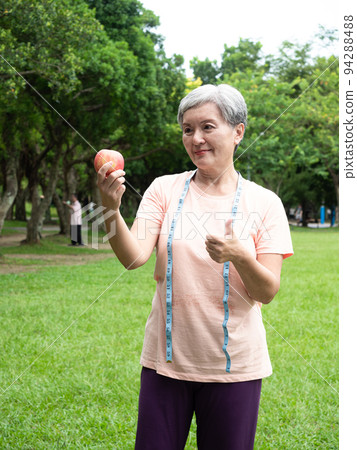 Portrait of happy senior adult elderly asia woman smiling standing with measuring tape and holding apple in the park. 94288488