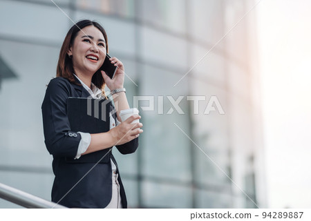 Young Asian businesswoman talking on phone and sitting in airport before business trip. Beautiful woman passenger has mobile call and discusses something with smile, holds coffee in hand Young Asian businesswoman talking on phone and sitting in airport before business trip. Beautiful woman passenger has mobile call and discusses something with smile, holds coffee in hand 94289887