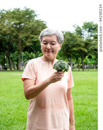 Happy senior asian woman smiling and standing with organic fruits and vegetables in hands enjoying nature fresh air in park, suitable for elderly healthy eating or vegetarian food 94290234