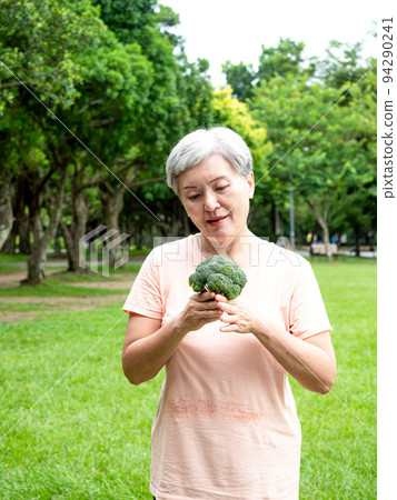 快樂的樂齡亞洲婦女微笑著站著,手上拿著有機蔬果,享受公園裡的大自然新鮮空氣,適用於老人健康飲食或素食 94290241