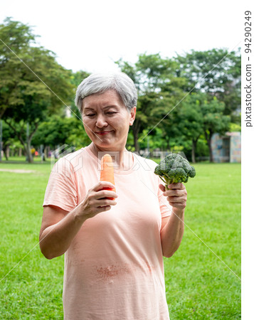 Happy senior asian woman smiling and standing with organic fruits and vegetables in hands enjoying nature fresh air in park, suitable for elderly healthy eating or vegetarian food 94290249