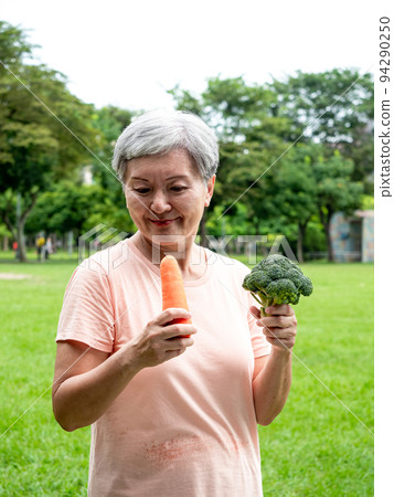 快樂的樂齡亞洲婦女微笑著站著,手上拿著有機蔬果,享受公園裡的大自然新鮮空氣,適用於老人健康飲食或素食 94290250
