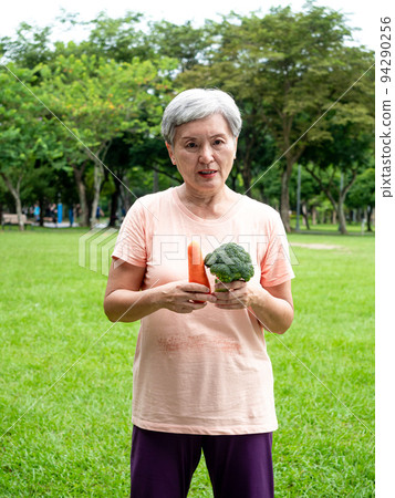 Happy senior asian woman smiling and standing with organic fruits and vegetables in hands enjoying nature fresh air in park, suitable for elderly healthy eating or vegetarian food 94290256