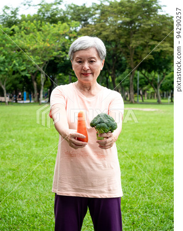 Happy senior asian woman smiling and standing with organic fruits and vegetables in hands enjoying nature fresh air in park, suitable for elderly healthy eating or vegetarian food 94290257