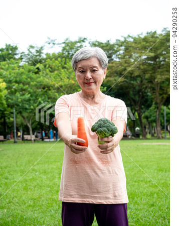 Happy senior asian woman smiling and standing with organic fruits and vegetables in hands enjoying nature fresh air in park, suitable for elderly healthy eating or vegetarian food 94290258