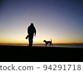 A person walking with a dog by the sea at sunset 94291718