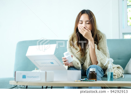 Young Asian woman sick, tired, she sitting and resting on blue fabric sofa and blanket after takes medicines pills in living room at her home. Woman use the phamacy first aid kit box from delivery Young Asian woman sick, tired, she sitting and resting on blue fabric sofa and blanket after takes medicines pills in living room at her home. Woman use the phamacy first aid kit box from delivery 94292913