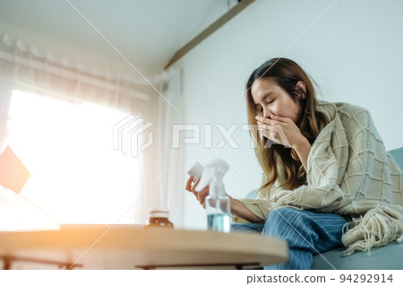 Young Asian woman sick, tired, she sitting and resting on blue fabric sofa and blanket after takes medicines pills in living room at her home. Woman use the phamacy first aid kit box from delivery 94292914