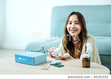 Young Asian woman sick, tired, she sitting and resting on blue fabric sofa and blanket after takes medicines pills in living room at her home. Woman use the phamacy first aid kit box from delivery Young Asian woman sick, tired, she sitting and resting on blue fabric sofa and blanket after takes medicines pills in living room at her home. Woman use the phamacy first aid kit box from delivery 94292916
