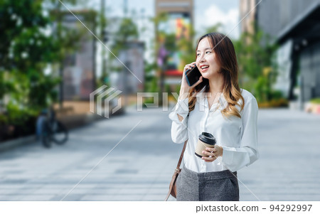 Smiling Asian curly business woman wearing trendy walks down the central city street and uses her phone. Pretty summer woman in white shirt walks down the street looking at her mobile phone 94292997