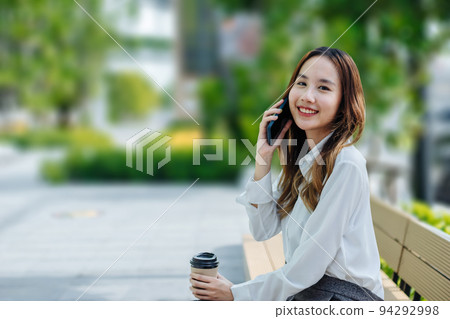 Smiling Asian curly business woman wearing trendy walks down the central city street and uses her phone. Pretty summer woman in white shirt walks down the street looking at her mobile phone 94292998