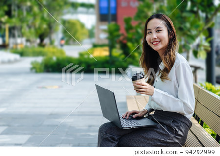 Smiling Asian curly business woman wearing trendy sitting in the central city street and uses her phone and laptop. Pretty summer woman in white shirt walks down the street looking at her mobile phone Smiling Asian curly business woman wearing trendy sitting in the central city street and uses her phone and laptop. Pretty summer woman in white shirt walks down the street looking at her mobile phone 94292999