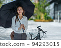 Young Asian businesswoman holding umbrella looking the watch and standing on a city street with bicycle looking at phone. 94293015