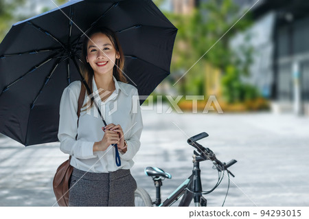 Young Asian businesswoman holding umbrella looking the watch and standing on a city street with bicycle looking at phone. 94293015