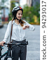Young Asian businesswoman wearing helmet and standing on a city street with bicycle looking at the watch. 94293017