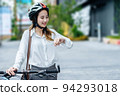 Young Asian businesswoman wearing helmet and standing on a city street with bicycle looking at sky or watch. 94293018