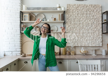 Happy young beautiful woman in glasses and green shirt dancing and singing at home in the kitchen. Resting from homework, relaxed, enjoying time alone Happy young beautiful woman in glasses and green shirt dancing and singing at home in the kitchen. Resting from homework, relaxed, enjoying time alone 94296658
