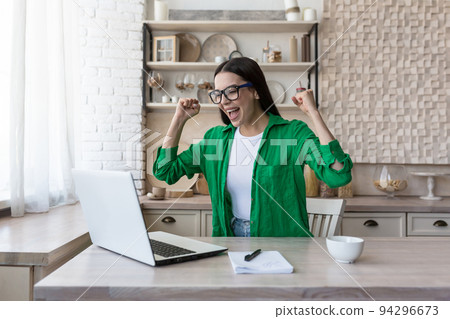 Portrait of happy young business woman in glasses and green jacket. She sits at the table at home, works with a laptop, is happy, raised her hands up, received good news, smiles. 94296673