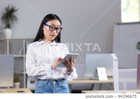 Portrait of young beautiful Asian female student in glasses, white shirt and jeans. Standing in the office, holding a tablet in his hands, studying, typing Portrait of young beautiful Asian female student in glasses, white shirt and jeans. Standing in the office, holding a tablet in his hands, studying, typing 94296675