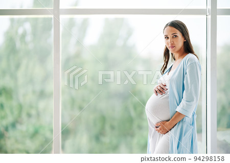 Portrait of young pregnant woman standing near window holding hands on her belly at modern home Portrait of young pregnant woman standing near window holding hands on her belly at modern home 94298158