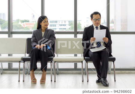 Young woman look and smile at a man sitting further away. Asian business people waiting for job interview by sitting spaced apart. Concept of new normal office during coronavirus pandemic outbreak Young woman look and smile at a man sitting further away. Asian business people waiting for job interview by sitting spaced apart. Concept of new normal office during coronavirus pandemic outbreak 94298602