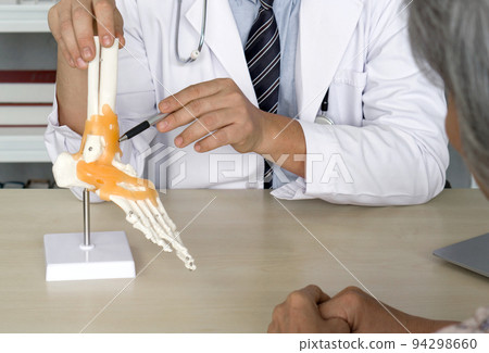 Orthopedic Surgeon in white gown and stethoscope pointing at human skeleton foot ankle bone joint anatomy model, present to the elderly patient about treatment of the ankle with surgery. 94298660