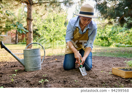 A woman farmer, sitting near a wetering can in the eco field, using a garden shovel, digging a hole in the loosen fertile ground for planting vegetables in the open field. The start of planting season A woman farmer, sitting near a wetering can in the eco field, using a garden shovel, digging a hole in the loosen fertile ground for planting vegetables in the open field. The start of planting season 94299568