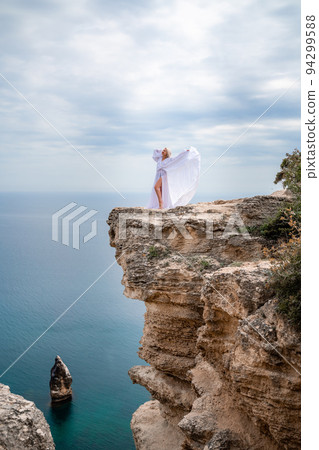 Blonde with long hair on a sunny seashore in a white flowing dress, rear view, silk fabric waving in the wind. Against the backdrop of the blue sky and mountains on the seashore. 94299588