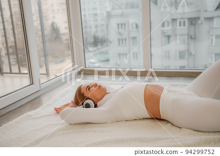 Side view portrait of relaxed woman listening to music with headphones lying on carpet at home. She is dressed in a white tracksuit. Side view portrait of relaxed woman listening to music with headphones lying on carpet at home. She is dressed in a white tracksuit. 94299752