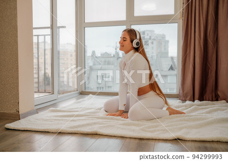 Side view portrait of relaxed woman listening to music with headphones lying on carpet at home. She is dressed in a white tracksuit. Side view portrait of relaxed woman listening to music with headphones lying on carpet at home. She is dressed in a white tracksuit. 94299993