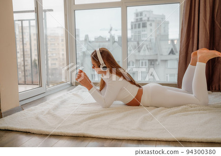 Side view portrait of relaxed woman listening to music with headphones lying on carpet at home. She is dressed in a white tracksuit. 94300380