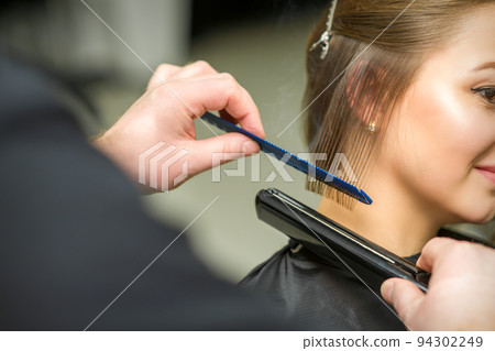 Hairstylist is straightening short hair of young brunette woman with a flat iron in a hairdresser salon, close up 94302249