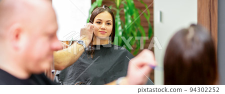 Reflection in the mirror of the young caucasian woman sitting and receiving haircut by male hairdresser at hairdresser salon Reflection in the mirror of the young caucasian woman sitting and receiving haircut by male hairdresser at hairdresser salon 94302252