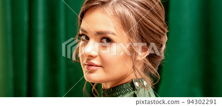 The fashionable young woman. Portrait of the beautiful female model with long hair and makeup with closed eyes. Beauty young woman with a brown curly hairstyle on the background of a green curtain The fashionable young woman. Portrait of the beautiful female model with long hair and makeup with closed eyes. Beauty young woman with a brown curly hairstyle on the background of a green curtain 94302291