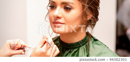 Makeup in the process. The hand of the makeup artist applies pink gloss lipstick on the lips of the beautiful face of the young caucasian woman in a beauty salon Makeup in the process. The hand of the makeup artist applies pink gloss lipstick on the lips of the beautiful face of the young caucasian woman in a beauty salon 94302301