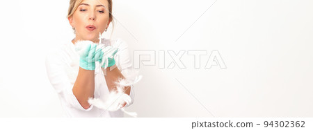 Young beautiful woman beautician in protective green gloves standing and blowing on feathers over white wall background 94302362