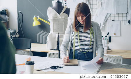 Young pretty woman is drawing in notepad looking at clothing sketch while working in her tailoring studio. Drawings hanging on wall, bright sewing items are visible. 94303037