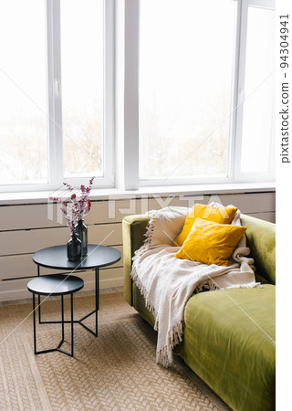 A green sofa with yellow pillows and a beige blanket, a coffee table with a vase and eucalyptus branches. Modern living room with large window 94304941