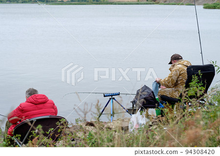 Two fishermen are sitting in chairs on a fishing trip on the shore of a reservoir, river, lake 94308420